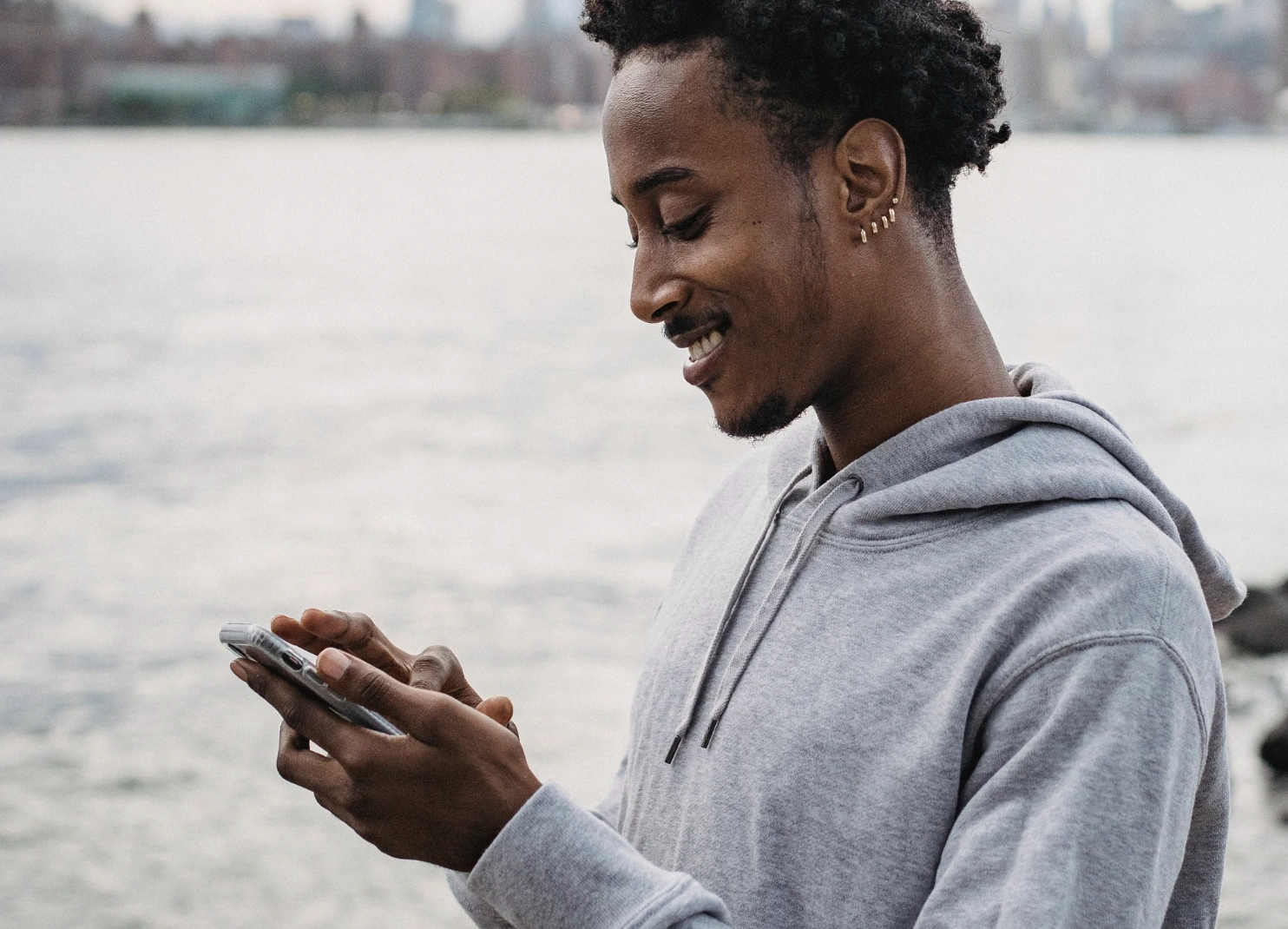 Smiling man in a grey hoodie using his phone by the waterfront