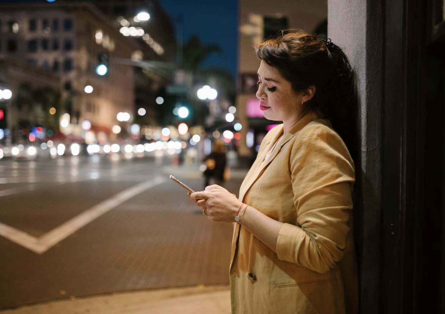 Woman in a beige blazer checking her phone on a city street at night